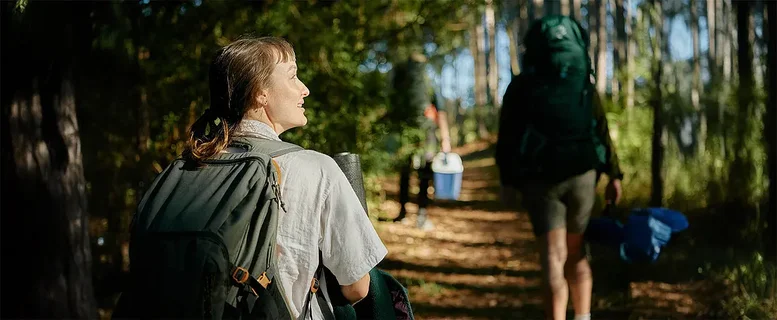 Eine Wanderin mit Rucksack lächelt auf einem Waldweg, während andere Wanderer folgen.