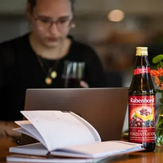 Eine Person arbeitet am Laptop mit einem Getränk und einem Notizbuch auf dem Tisch.