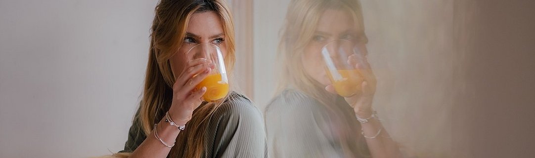 Frau mit langen Haaren trinkt aus einem Glas mit Orangensaft, Spiegelung im Hintergrund.