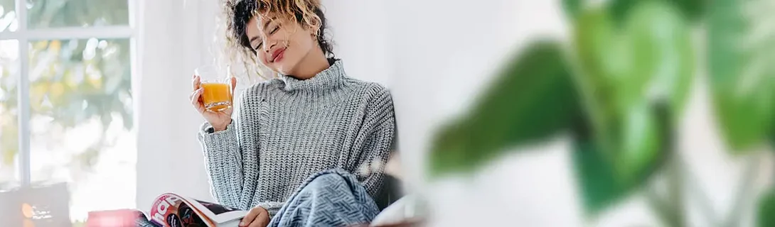 Junge Frau mit lockigem Haar sitzt entspannt und hält ein Glas Saft in der Hand.