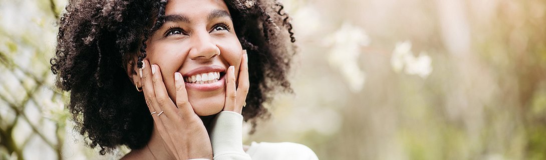 Junge Frau mit lockigem Haar lächelt und hält ihr Gesicht in den Händen.