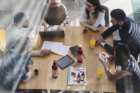 Sechs Personen sitzen an einem Tisch mit Laptops, Getränken und Dokumenten.